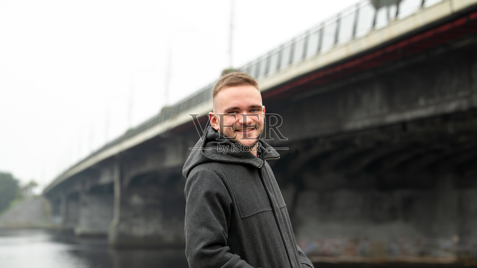 年轻男子自信地站在河边的桥下,微笑着穿着休闲服,享受着宁静的氛围,雾气弥漫的日子。照片摄影图片