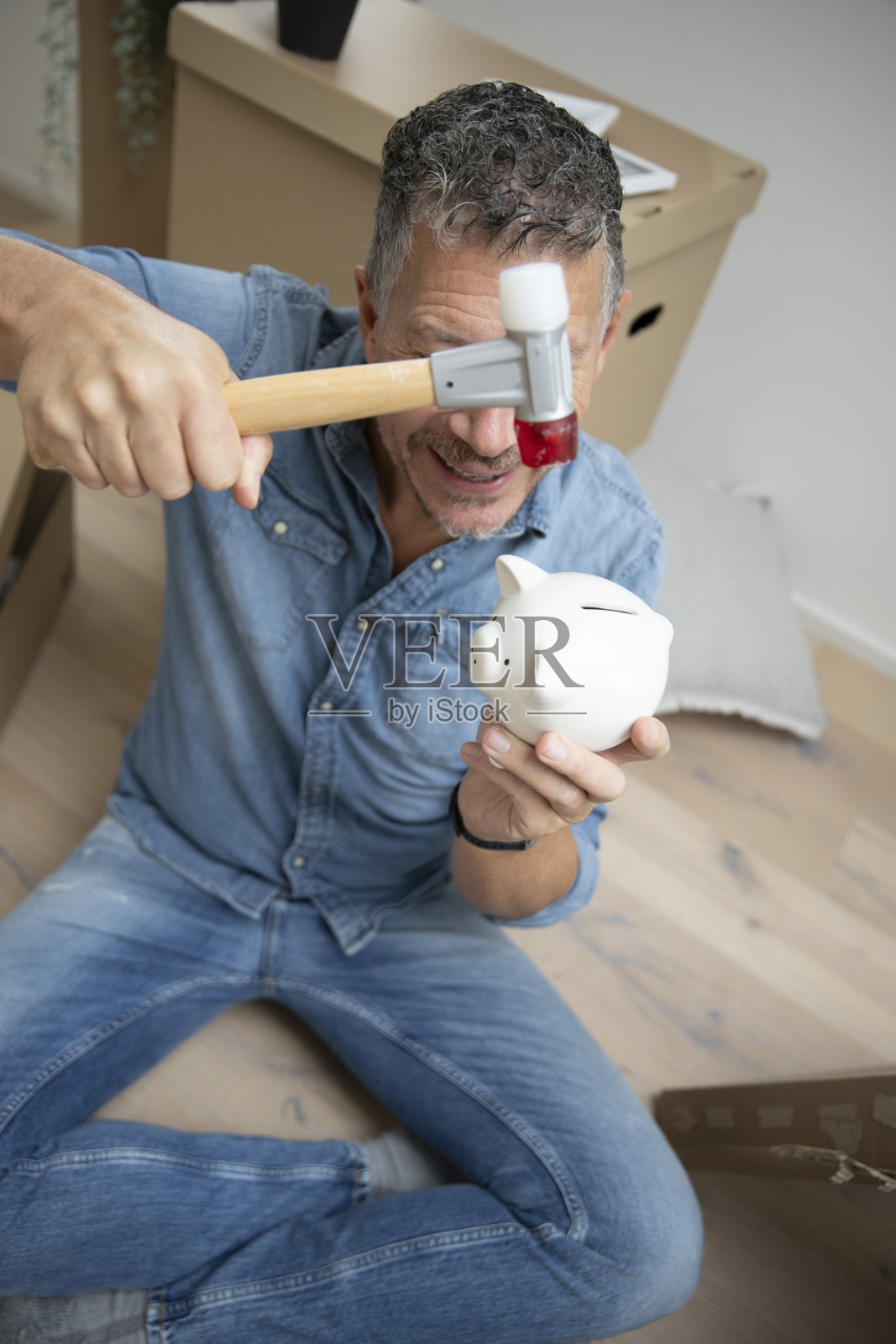 一位有着灰色头发的男子在他的新公寓、房子或阁楼里,周围有许多搬家箱子和一个储蓄罐。照片摄影图片