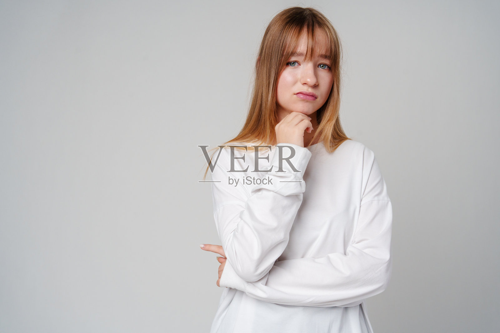 年轻女性穿着白色长袖衬衫,手托下巴,深思熟虑地摆姿势,背景简约。照片摄影图片