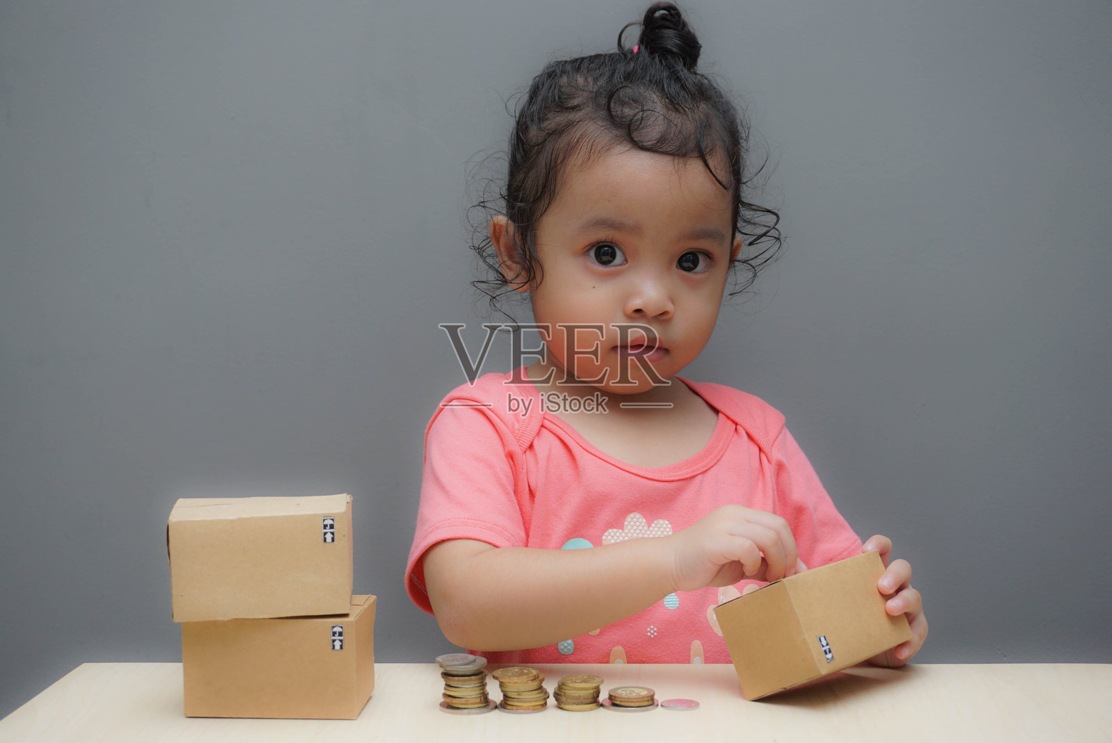 可爱女孩手持纸箱,桌上堆放着硬币,背景为灰色墙壁照片摄影图片