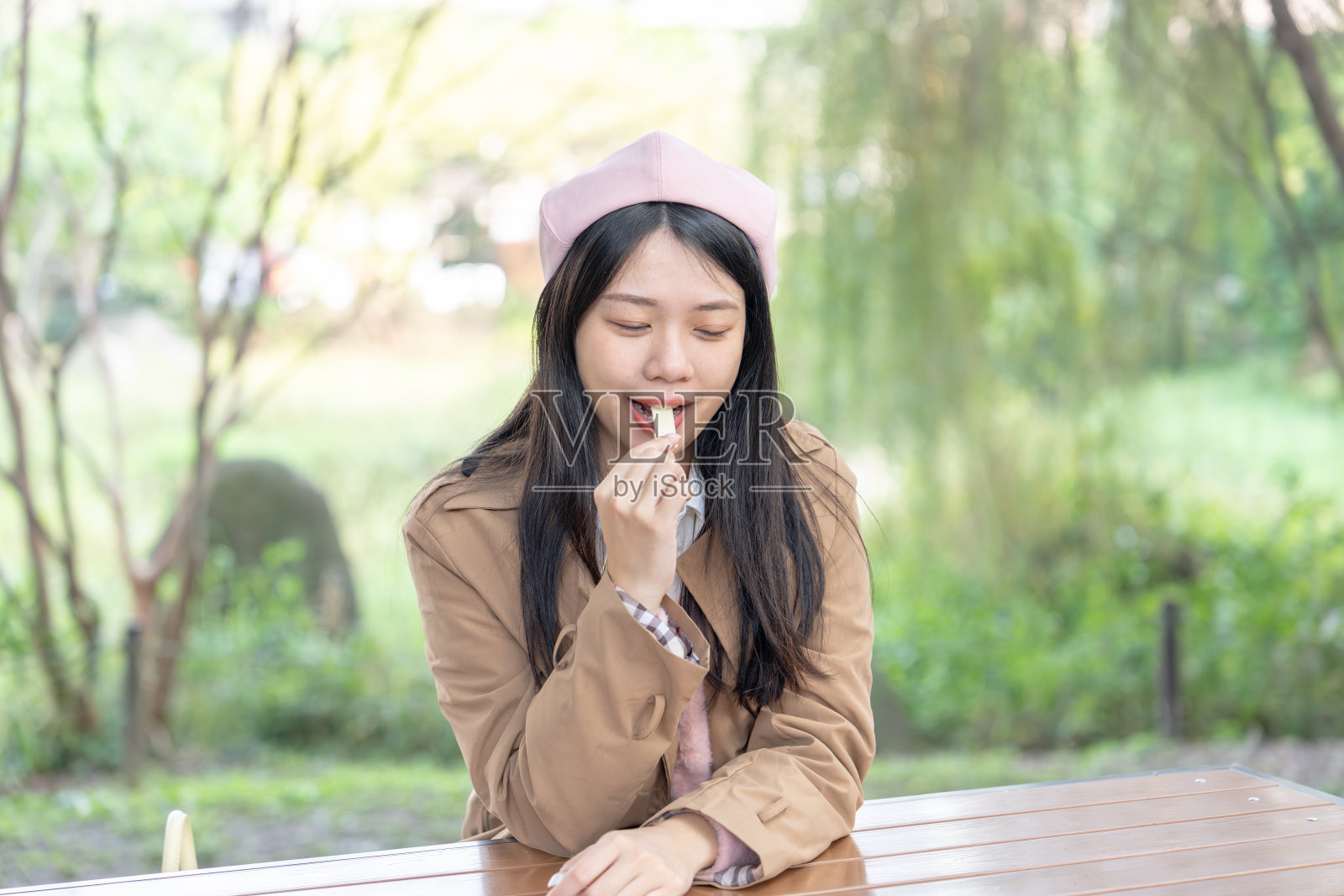 一位二十多岁的长发台湾女性穿着棕色风衣,坐在台北信义区公园的长椅上,冬季的1月30日中午享用巧克力。照片摄影图片