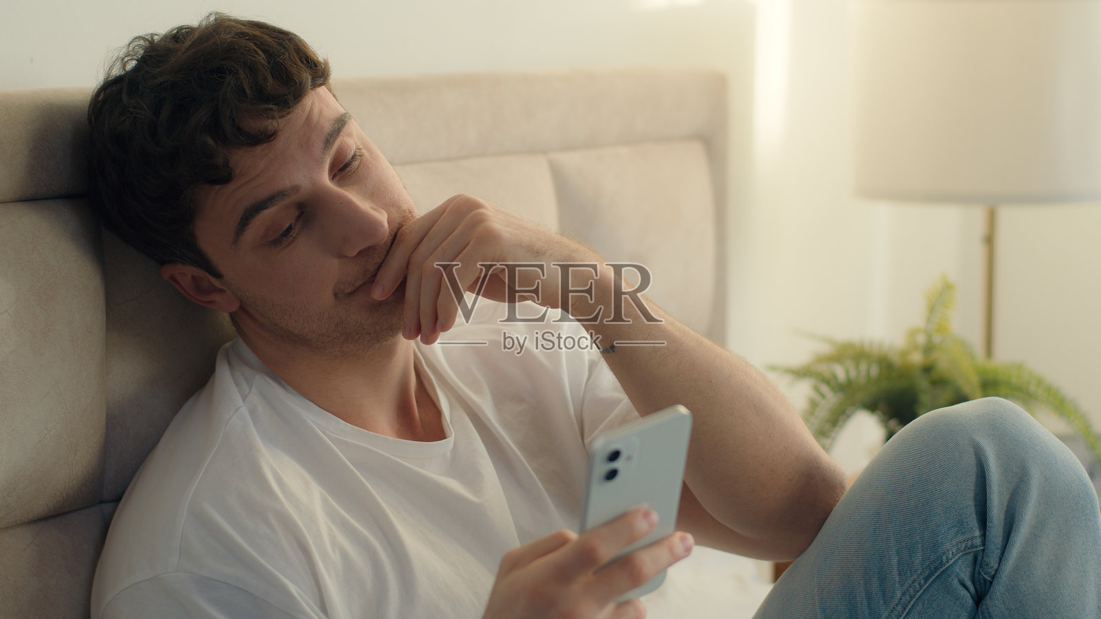 白人欧洲男性在卧室里感到疲惫,因过度工作而感到疲惫,手持手机在线工作,作为一名创业者,感到精疲力竭、困倦和沮丧。照片摄影图片