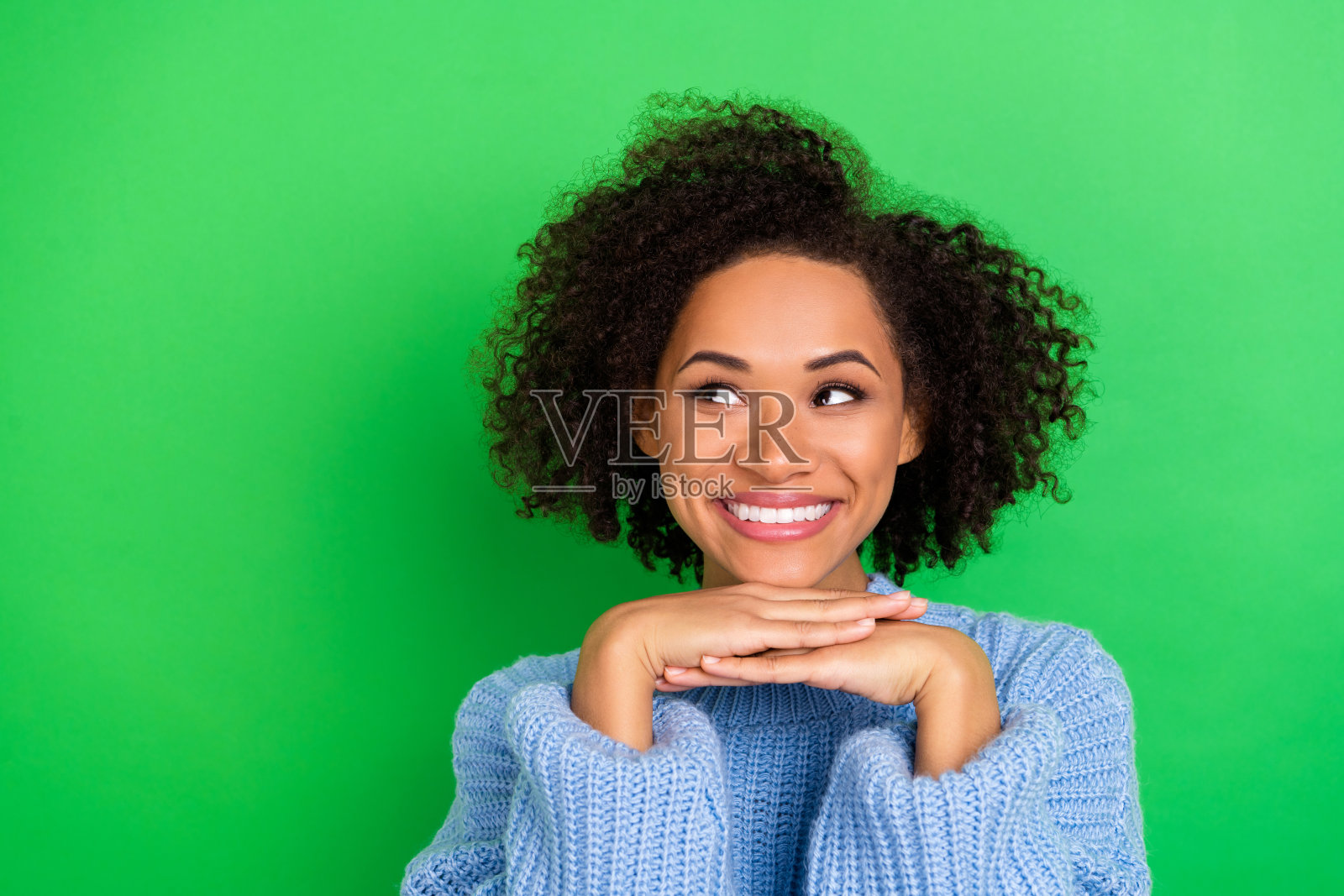 一位闪亮、可爱、笑容灿烂的女性,拥有波浪形发型,穿着蓝色毛衣,目光投向空旷的地方,背景为绿色。照片摄影图片