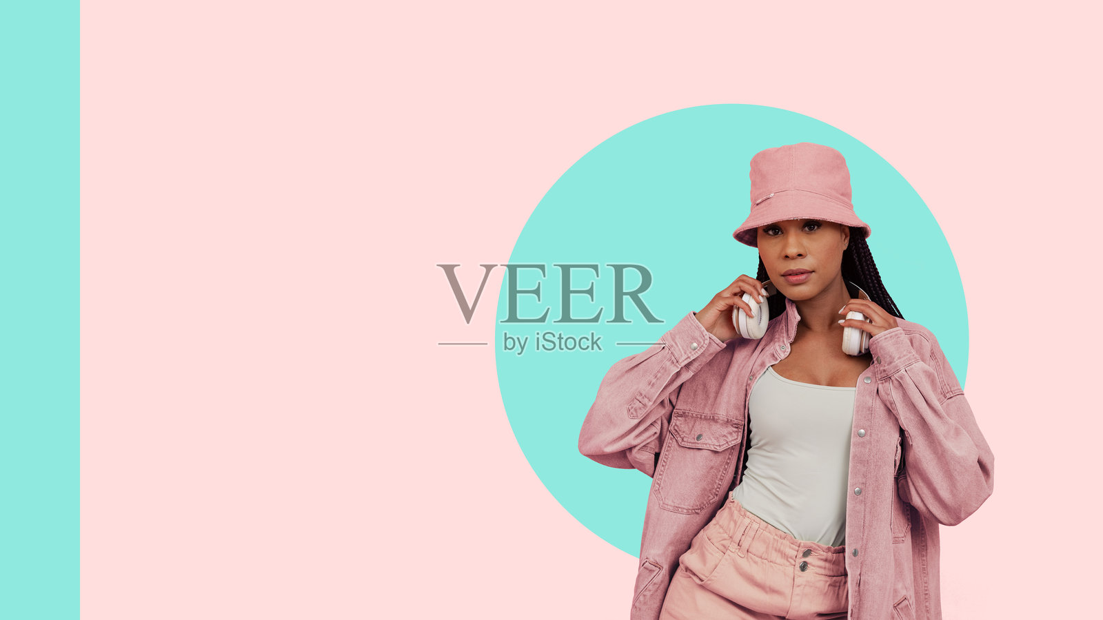 年轻女性穿着粉色休闲服和渔夫帽,手持耳机,背景为淡薄荷绿和桃色。照片摄影图片