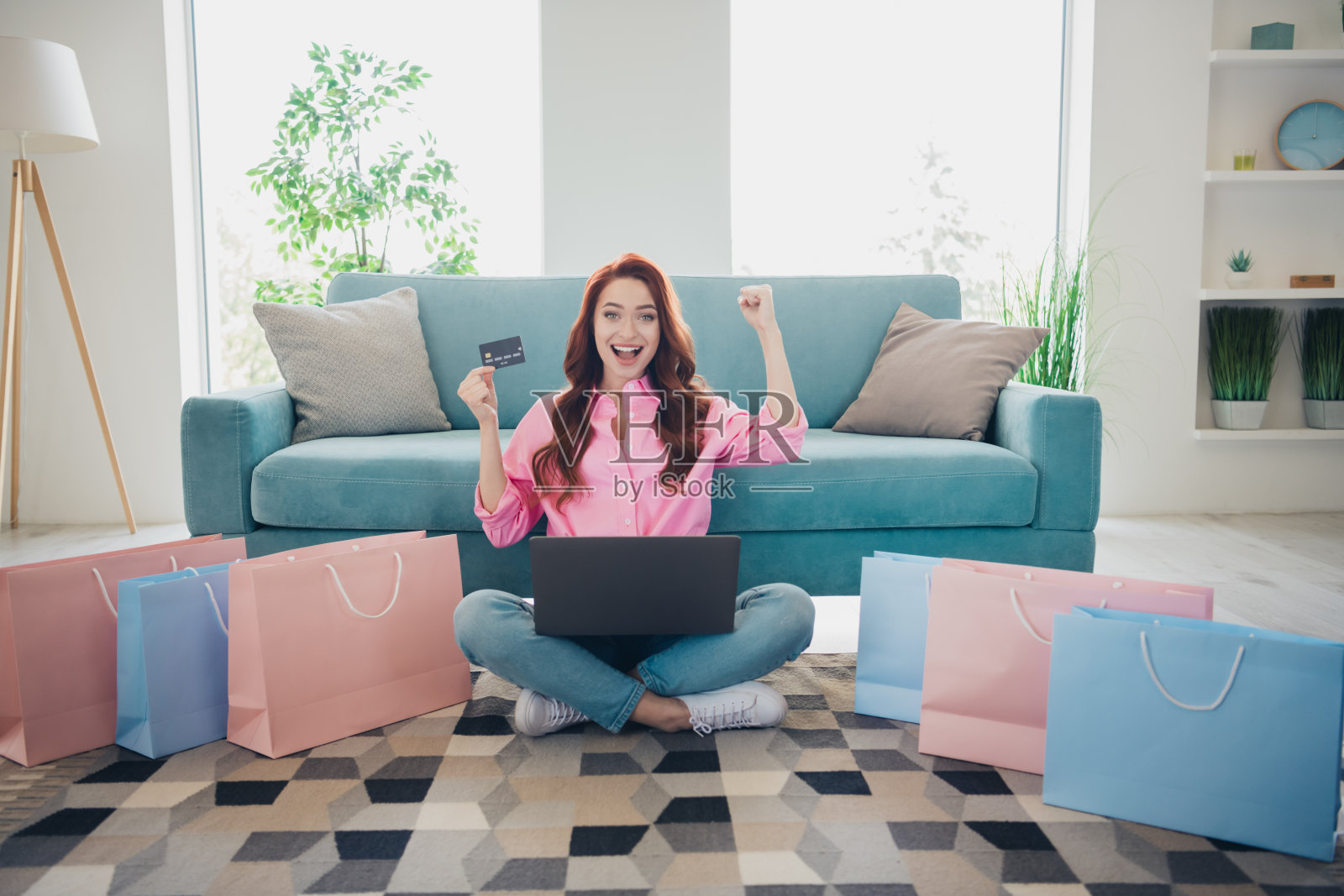 年轻漂亮的女性在现代明亮的公寓室内举起拳头,手里拿着信用卡和笔记本电脑,身边有购物袋。照片摄影图片