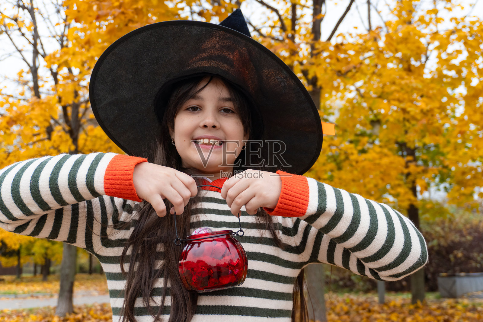 微笑的女孩打扮成女巫,手里拿着装有糖果的红色罐子,站在秋天的公园里照片摄影图片