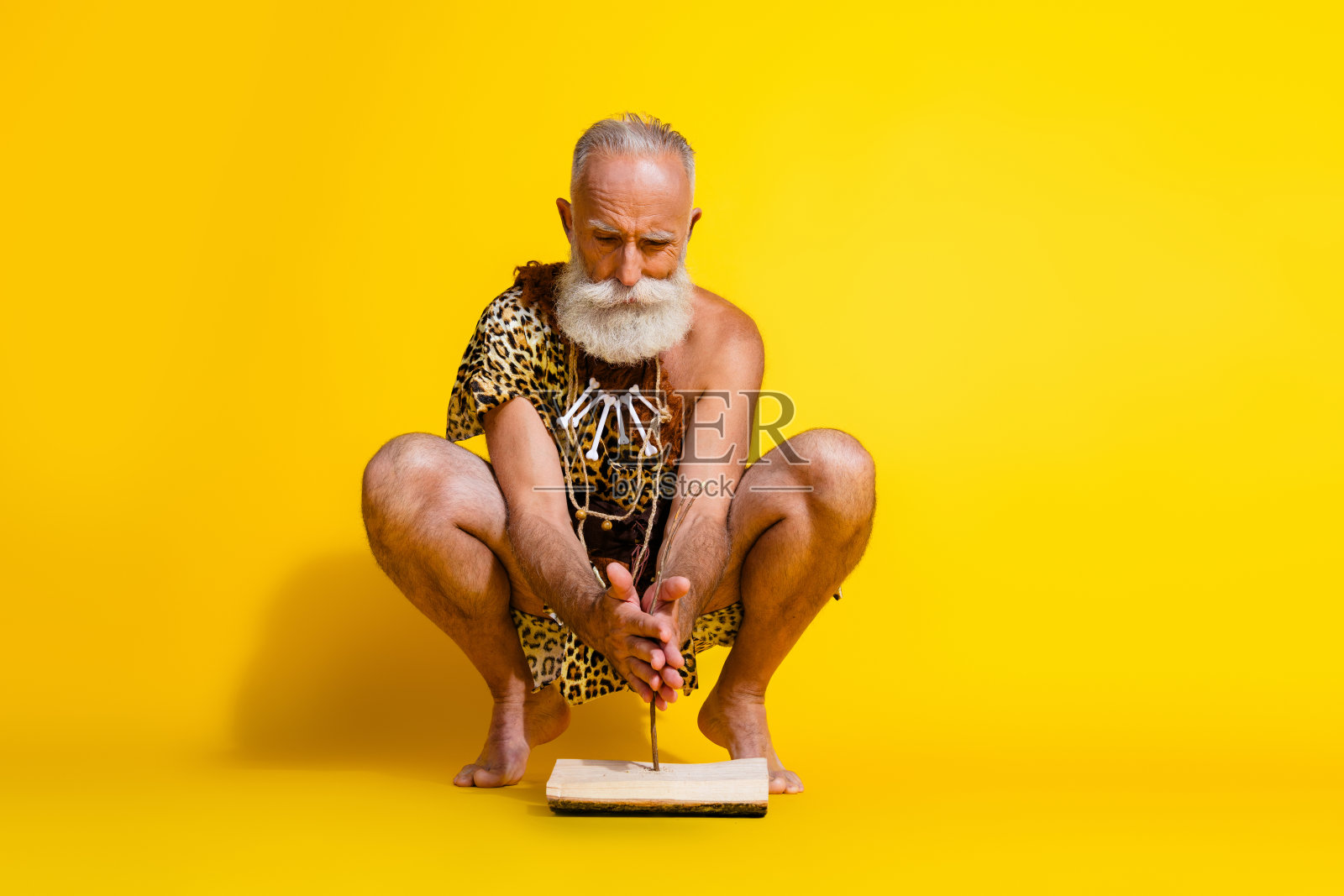 原始穴居人全身肖像,手持木棍生火,穿着豹纹皮肤服装,背景为黄色。照片摄影图片