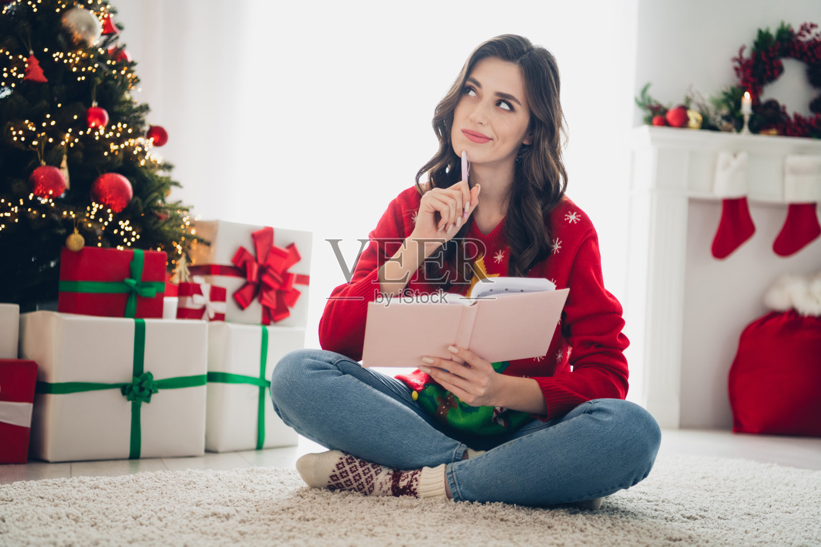 可爱的年轻女孩坐在地板上,手握铅笔和日记本,脑海中思考着,旁边是装饰有灯饰的圣诞树和堆放的礼物。照片摄影图片