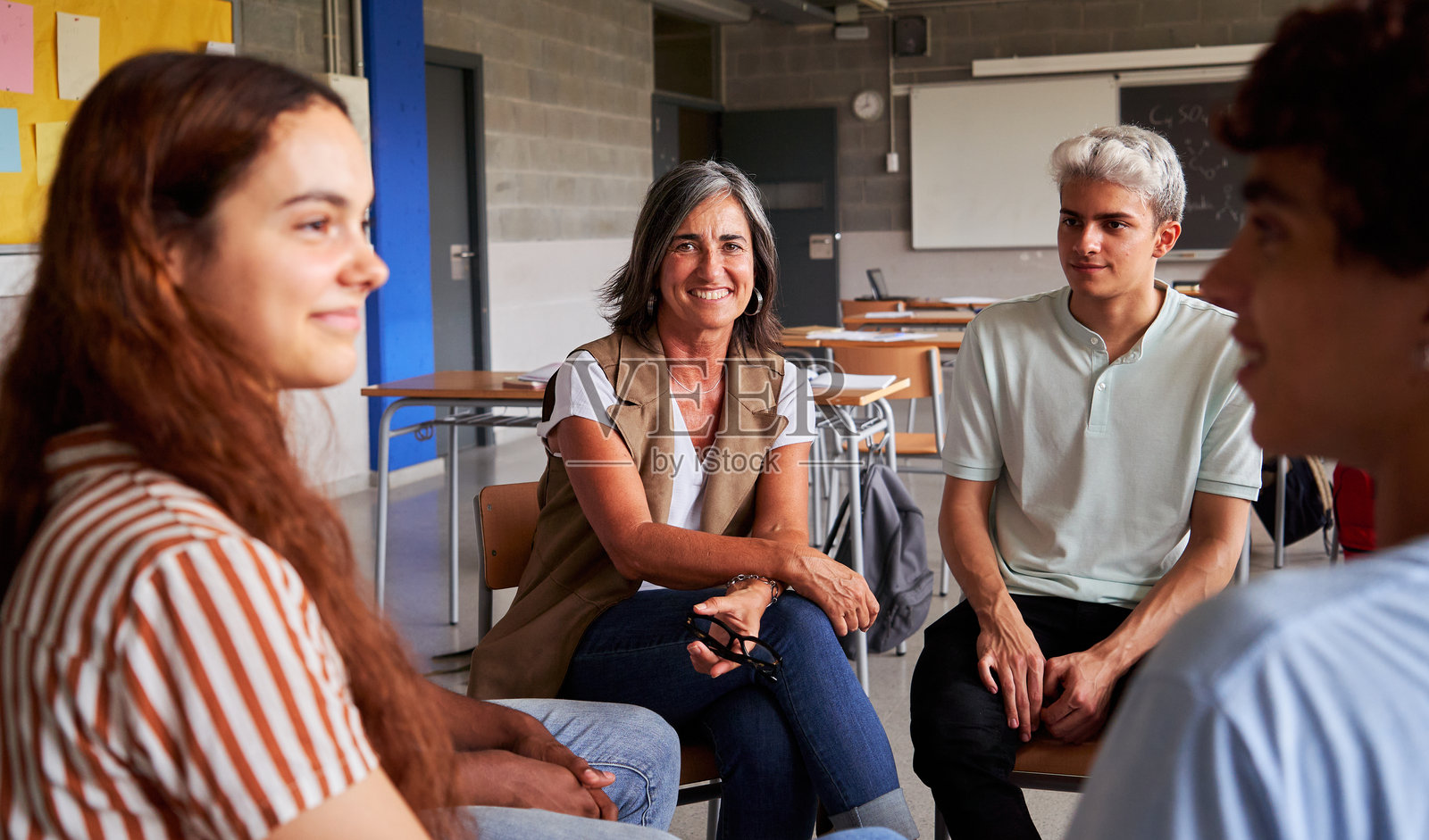一位成熟的女教师坐在大学生的圈子里,面带微笑地看着镜头。照片摄影图片