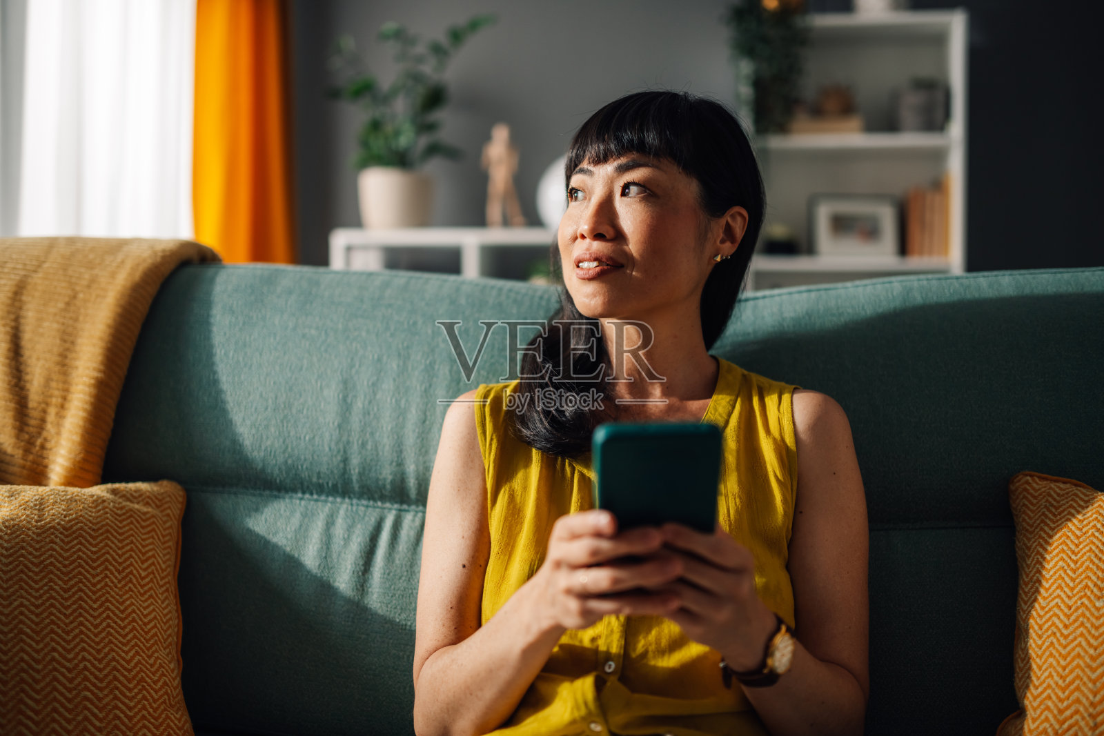 快乐的亚洲女性在阳光明媚的房间里,坐在绿色沙发上使用智能手机。照片摄影图片