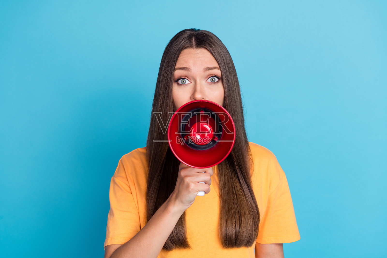害怕的漂亮女士穿着黄色衣服,手臂举着扩音器,宣布秘密消息,背景为蓝色。照片摄影图片