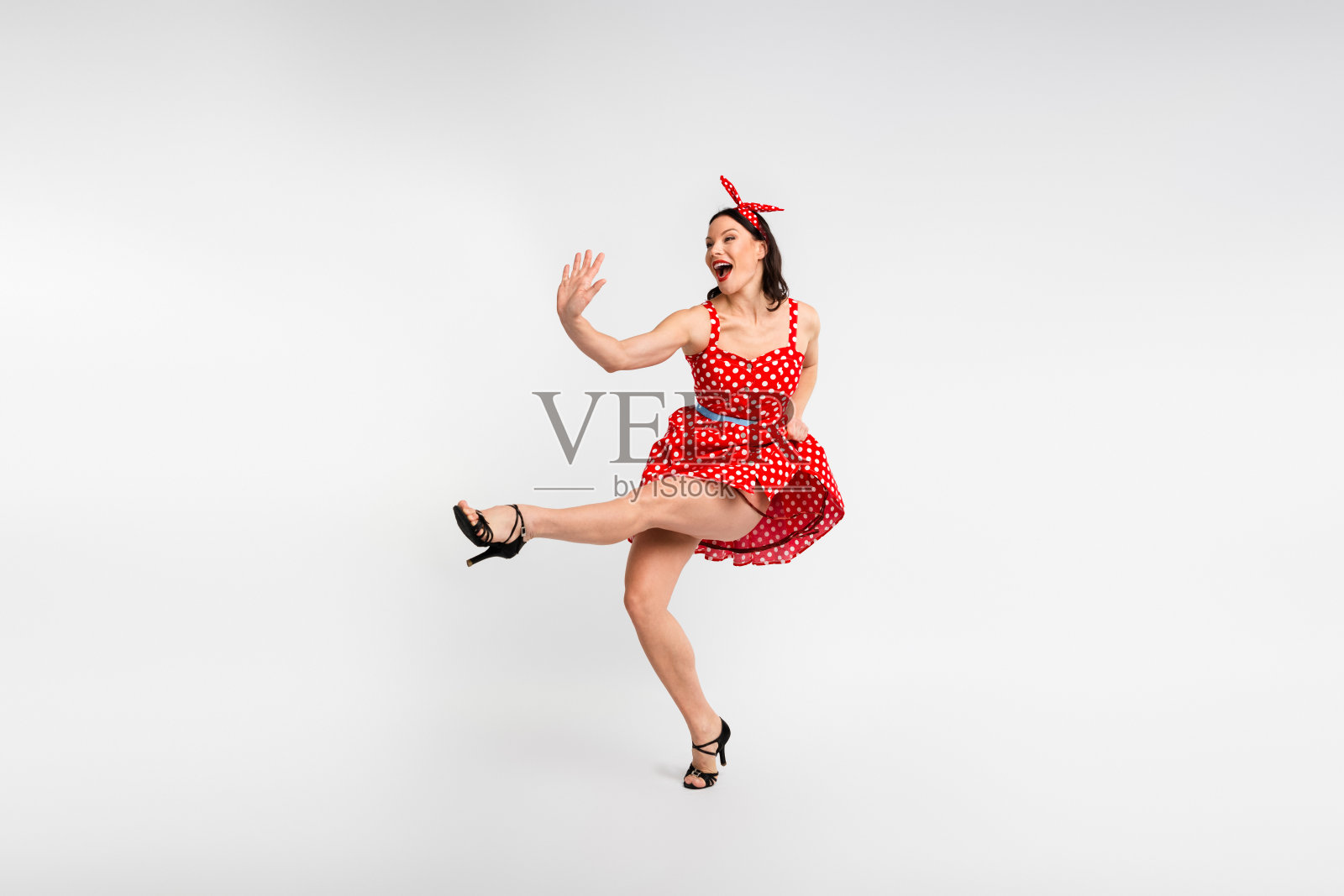 全身照,迷人的女士穿着红色复古风格的波点裙,开心地跳舞,抬起一条腿,背景为白色。照片摄影图片