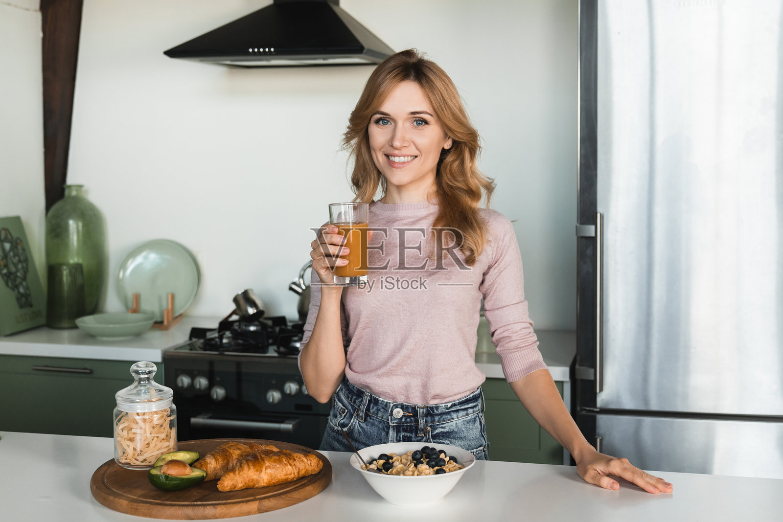 美丽的白人年轻女性,金发,穿着休闲服,站在现代厨房里,正在烹饪准备早餐,喝新鲜的果昔,面向镜头。照片摄影图片