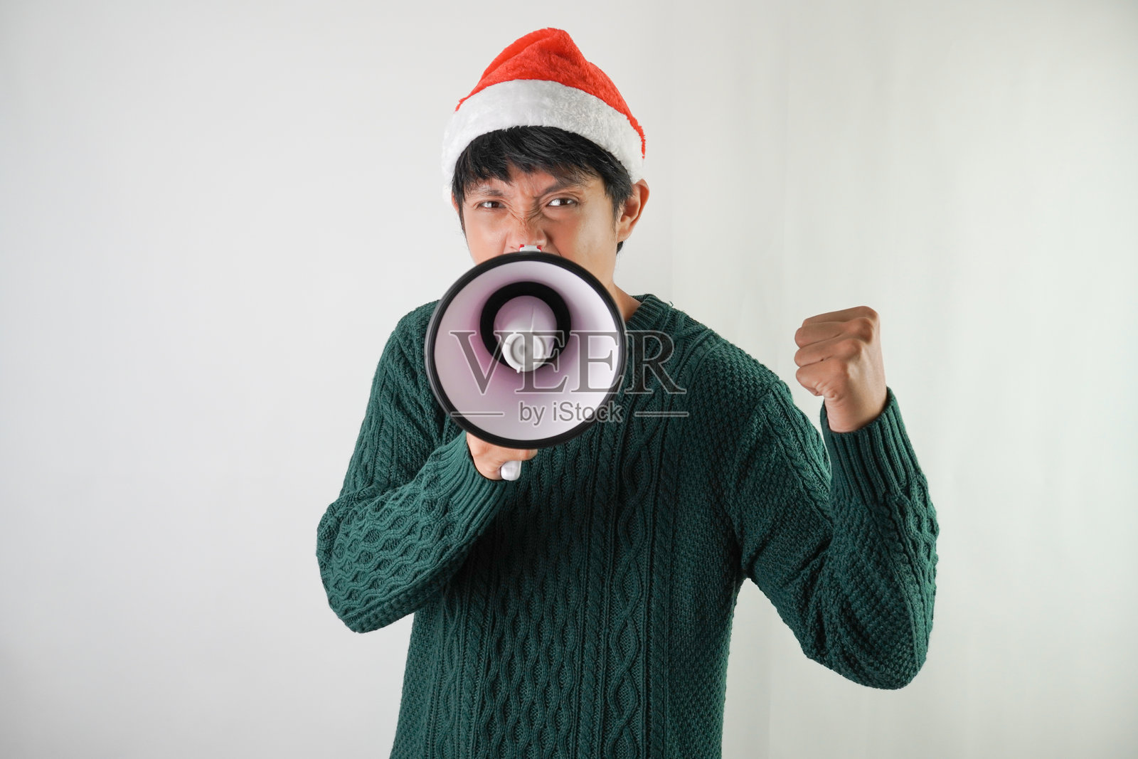 兴奋的年轻亚洲男子戴着圣诞老人帽,面带微笑,使用扩音器,背景为白色。适用于圣诞节和新年派对的概念。照片摄影图片