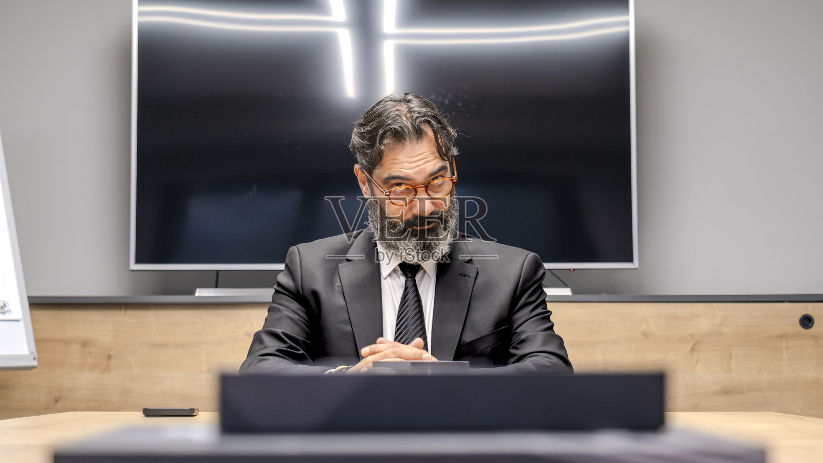 一位中年、深色头发、留着胡子的商人在办公室的会议桌旁坐着,面带紧张表情地看着他的员工。照片摄影图片