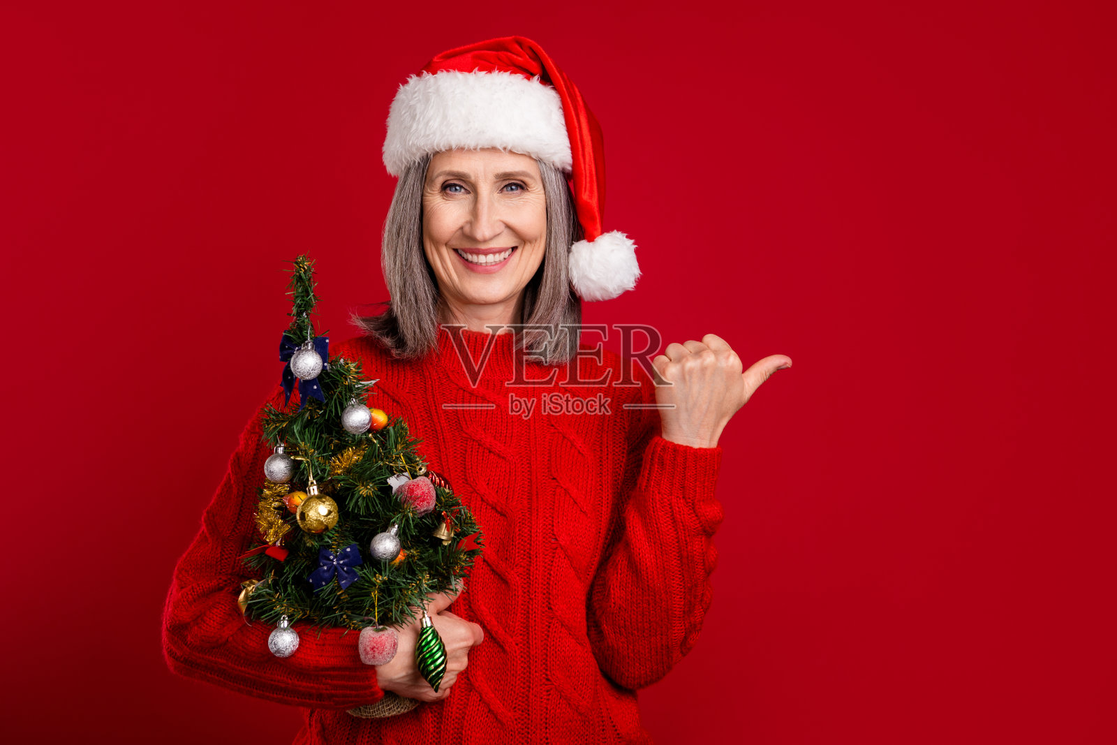 圣诞老人奶奶手持装饰树,指向空白区域,戴着帽子,穿着针织毛衣,背景为红色。照片摄影图片