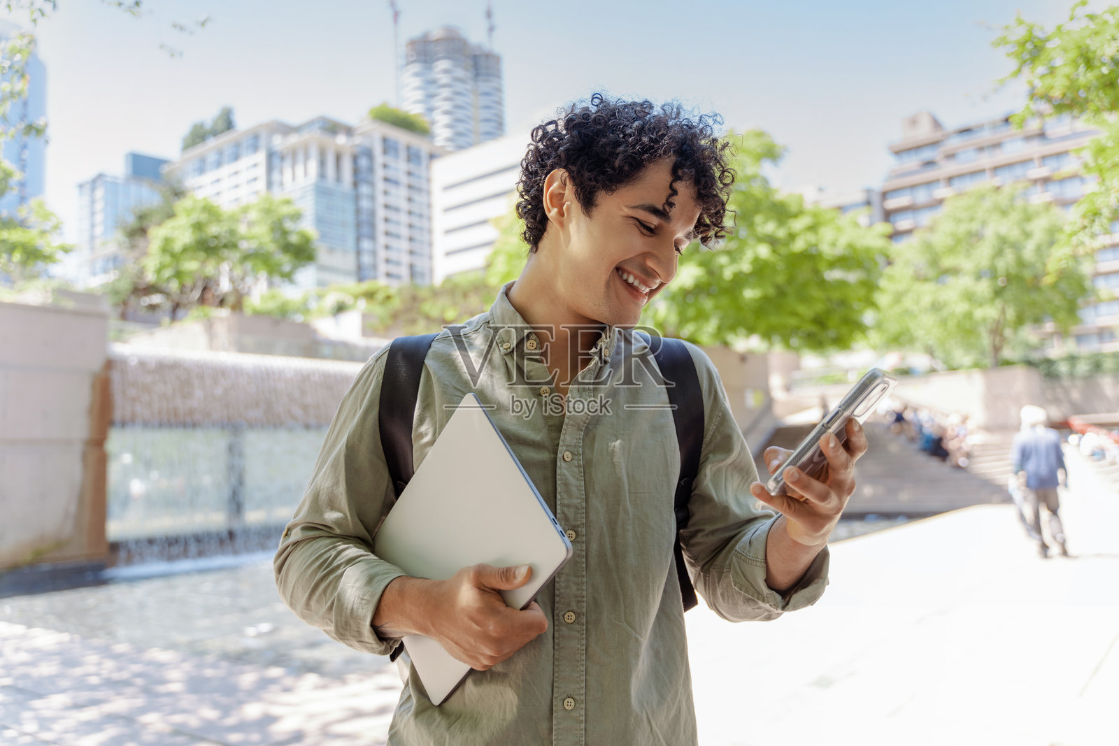 微笑的年轻男子在户外使用智能手机,手里拿着笔记本电脑照片摄影图片