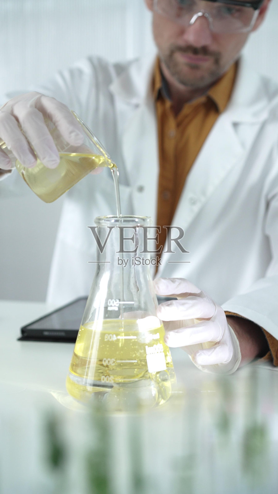 穿着实验室外套、白色手套和防护眼镜的男科学家正在实验室中将黄色油状液体从一个烧杯倒入另一个烧杯,旁边有显微镜,垂直特写视角。照片摄影图片