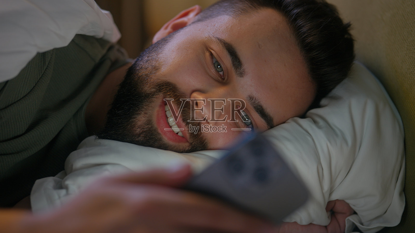 白人男性躺在床上使用手机,享受舒适的周末时光,微笑着放松,观看视频。照片摄影图片