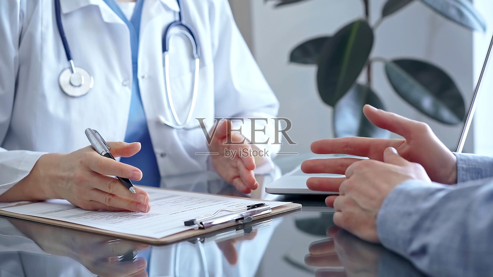穿着白色医师外套的女医生在诊所办公室咨询患者,两人坐在玻璃桌旁。医生在倾听并做笔记。医学概念照片摄影图片