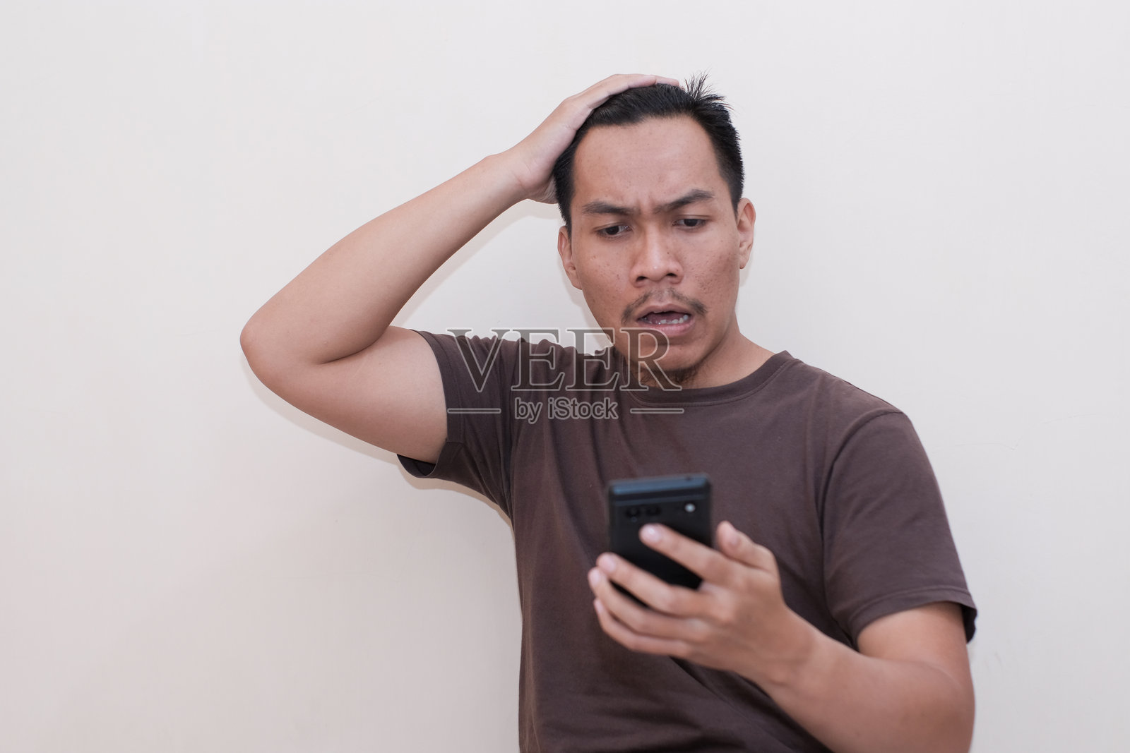 一位穿着棕色T恤的东南亚男子对手机中看到的内容感到震惊。照片摄影图片