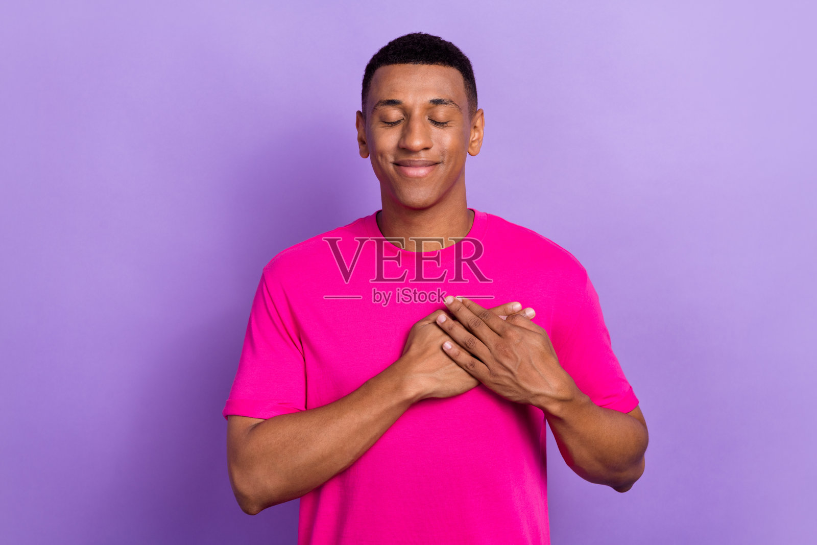 心情愉快、满意、感激的男士,短发,穿着粉色T恤,双眼闭合,双手放在心口,背景为紫色。照片摄影图片