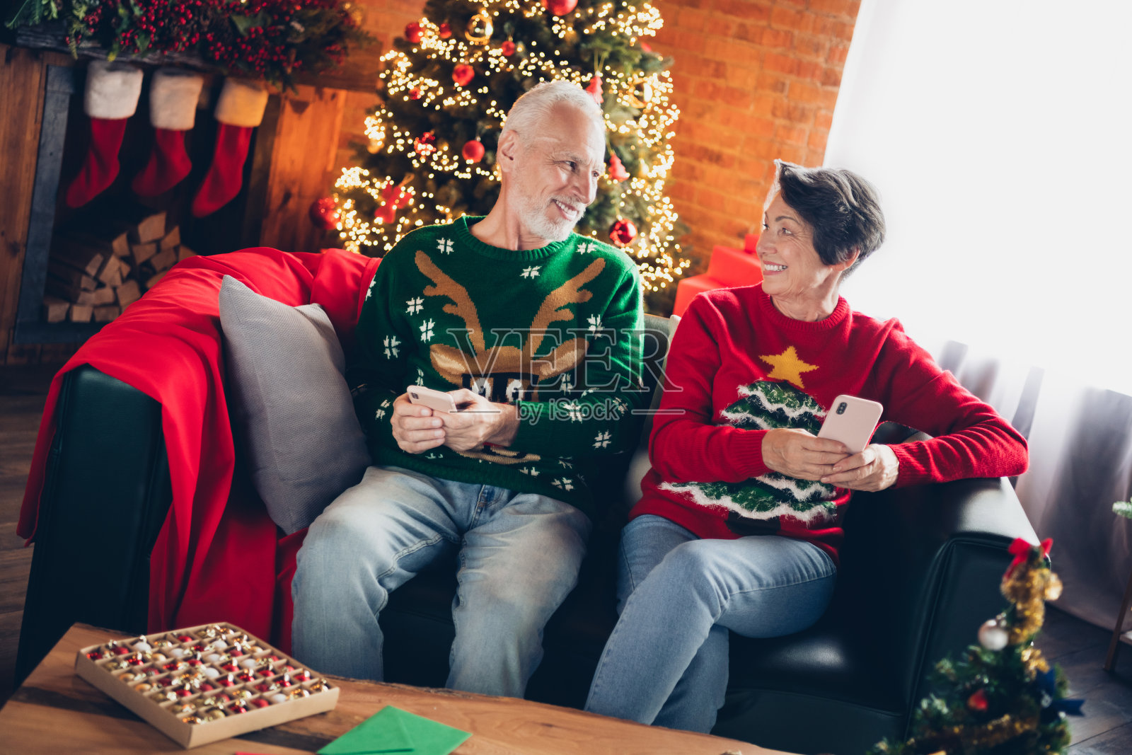 两位幽默积极的老年人坐在沙发上,手持电话,相互对视,室内有树木装饰。照片摄影图片