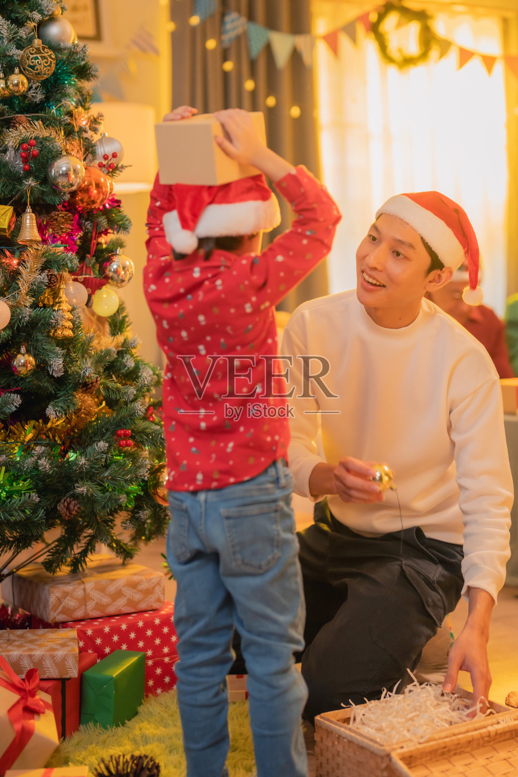 节日季节中捕捉到的快乐时刻,孩子穿着红色毛衣,举着礼物,身旁戴着圣诞老人帽子的男人微笑,周围是装饰华丽的圣诞树和节日装饰。照片摄影图片