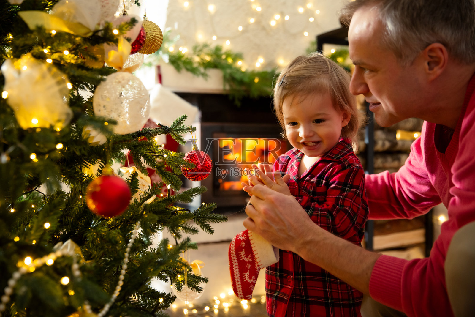 父亲和他兴奋的幼儿在家庆祝圣诞节。家人们一起快乐地装饰圣诞树。可爱的宝宝穿着红色格子睡衣。美丽的房子里有壁炉,地上摆满了包好的礼物。照片摄影图片