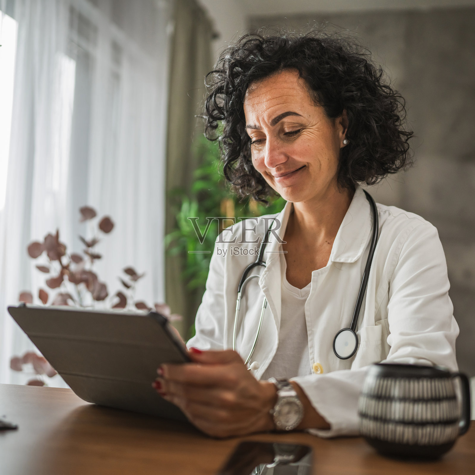 成熟的女性医生坐在桌子旁,使用数字平板电脑在家中。照片摄影图片