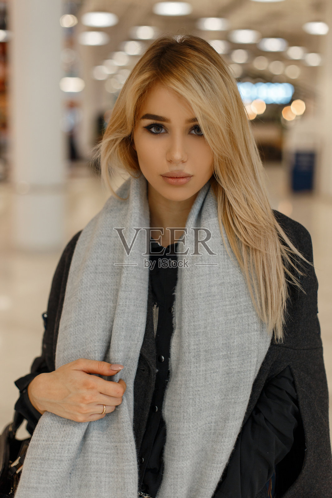 迷人的年轻金发美女,灰色眼,穿着时尚的灰色外套,搭配流行的围巾和手提包,站在明亮的商店里。优雅迷人的女孩模特在购物中心走动。照片摄影图片