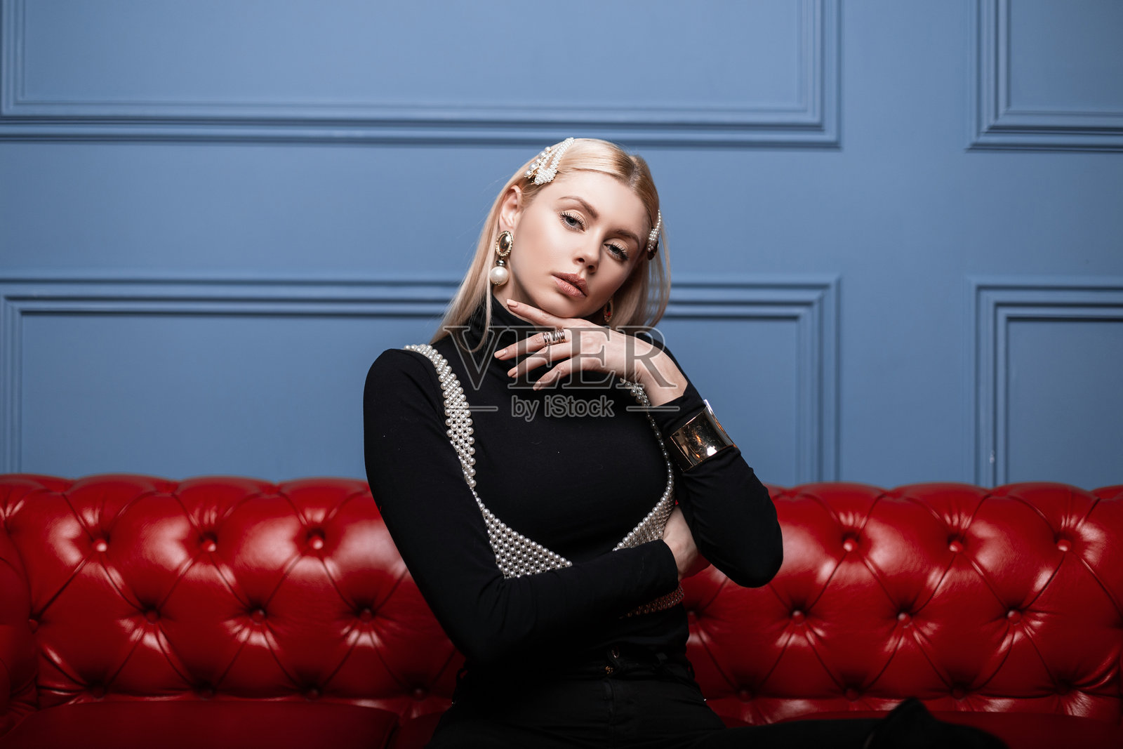 时尚的年轻女性,金发,佩戴优雅的珍珠配饰,身穿黑色时尚服装,搭配时尚的珍珠上衣,坐在房间里的红色皮沙发上摆姿势。现代女孩时尚模特室内。照片摄影图片