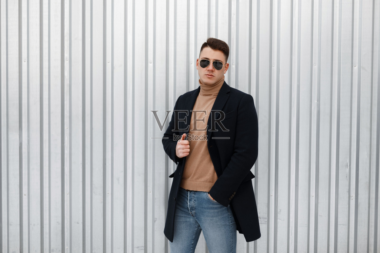 优雅的年轻时尚男士,穿着针织毛衣和时尚黑色外套,戴着太阳镜,搭配时尚牛仔裤和别致发型,站在金属墙旁。英俊的男孩。照片摄影图片