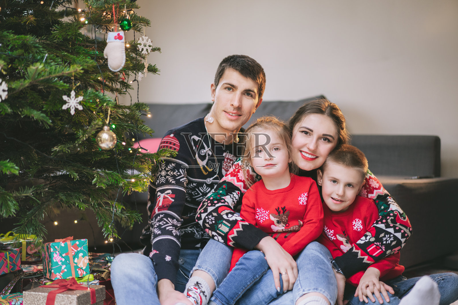 穿着节日毛衣的微笑家庭肖像,坐在家中的圣诞树下,周围是礼物。快乐的父母和孩子们依偎在一起,感到兴奋。家庭团聚时光。快乐的童年。照片摄影图片