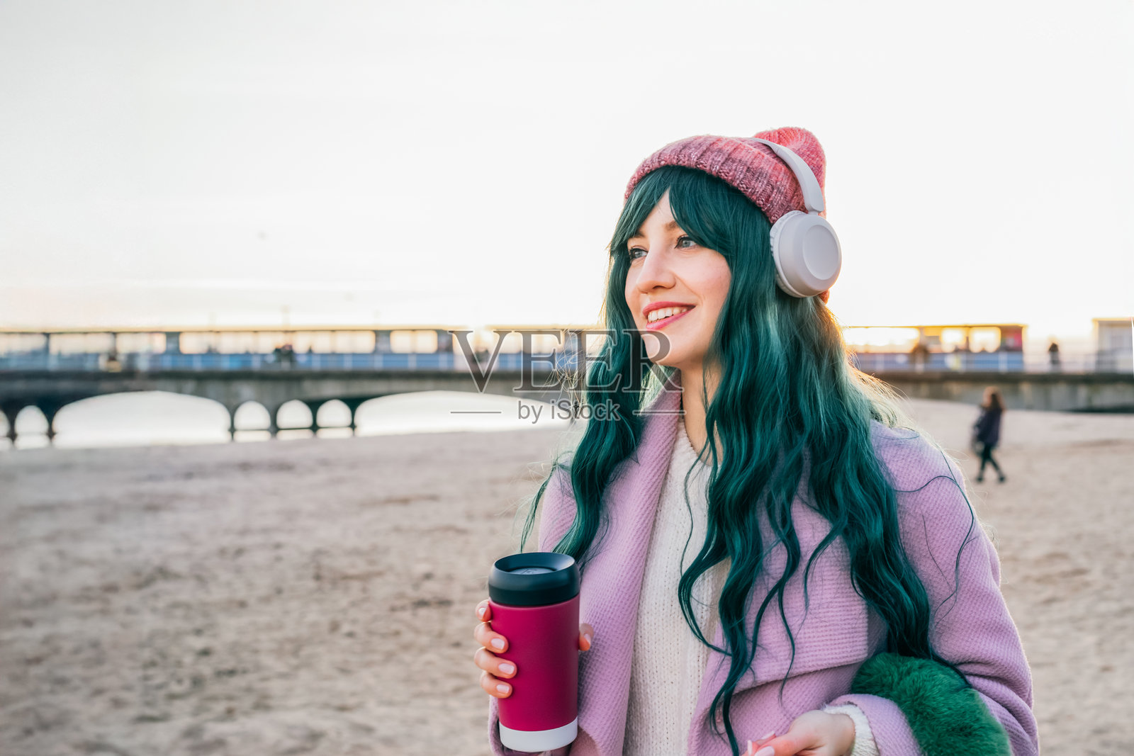 时尚的年轻微笑嬉皮士女性,染发,佩戴无线耳机,穿着粉色外套和帽子,手持可重复使用的咖啡杯,在海滩上散步。放松时刻。季节性时尚潮流。芭比风格。照片摄影图片