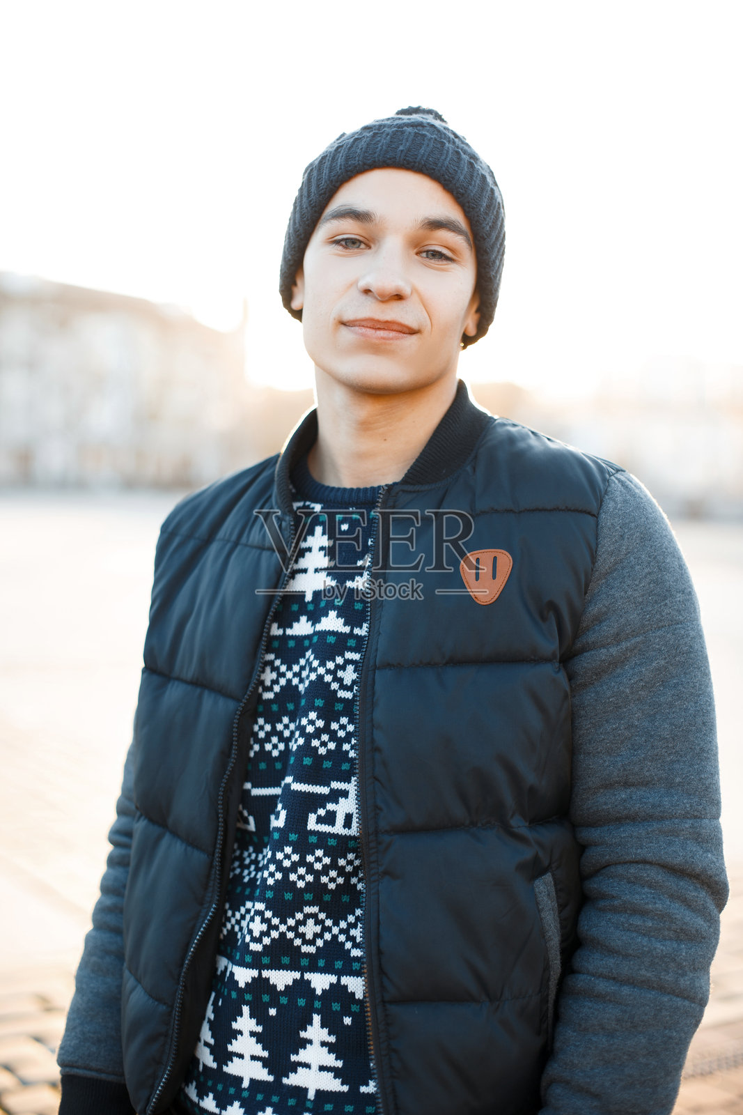 身穿时尚夹克、复古风格帽子和圣诞毛衣的帅气年轻男子,面带甜美微笑,站在城市中。时尚快乐的迷人男孩在散步。照片摄影图片