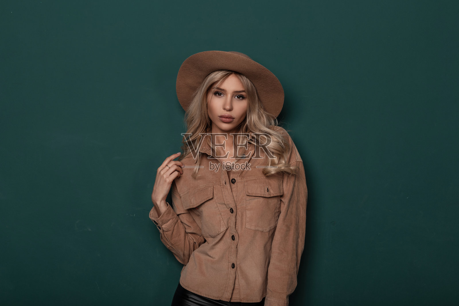 优雅的米色帽子、时尚衬衫、金色卷发的美丽迷人年轻女性站在工作室里,靠近绿色复古墙壁。迷人的时尚女孩。新款女装系列。照片摄影图片