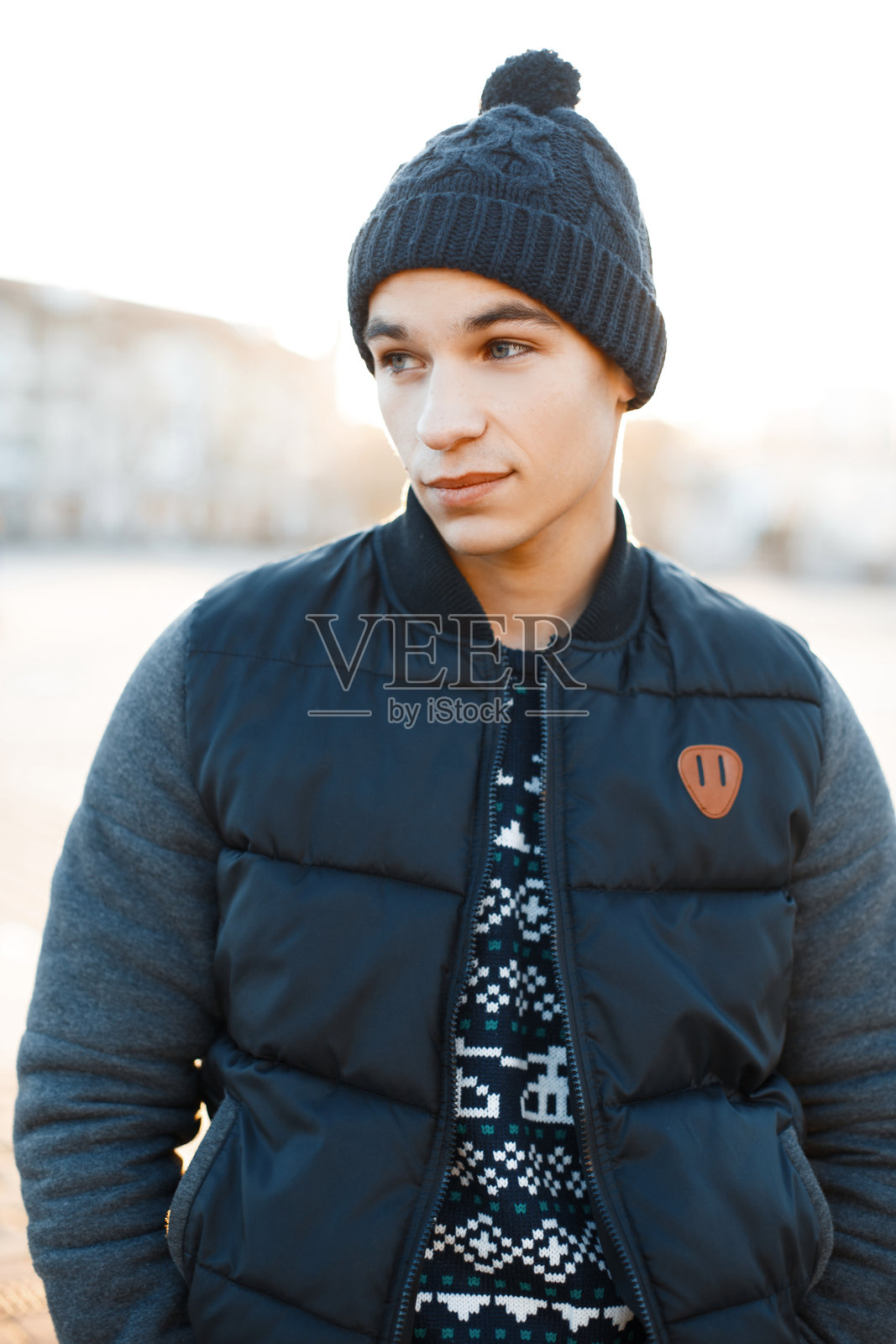 时尚迷人的年轻男子,戴着时尚的帽子,穿着蓝色复古针织毛衣和流行的温暖冬季外套,站在雪-covered城市的背景下,阳光明媚的冬日。英俊的男孩。照片摄影图片