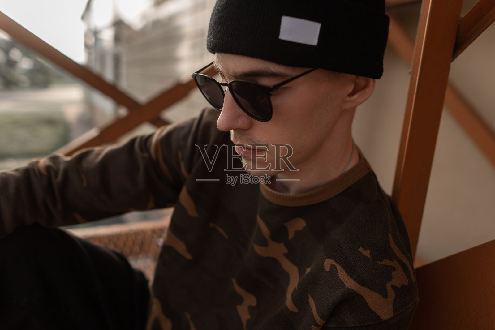 时尚的年轻嬉皮士男士,戴着流行的深色太阳镜,穿着时尚的针织帽和军装衬衫,站在户外的复古铁管旁。这个美丽的现代男孩在夏日里放松身心。照片摄影图片