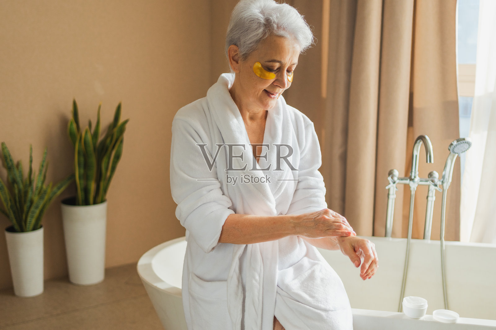 成熟的老年女性在手上涂抹保湿身体乳。老年女士在浴室里进行早晨日常护肤程序,涂抹滋养抗衰老面霜。美容护理水疗身体护肤。照片摄影图片