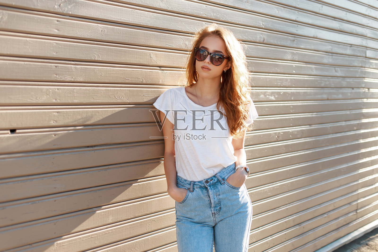 时尚的年轻嬉皮女孩,戴着时尚的太阳镜,穿着流行的白色T恤和复古牛仔裤,在阳光明媚的夏日里靠在木墙旁摆姿势。时尚女孩模特享受着春天的气息。照片摄影图片