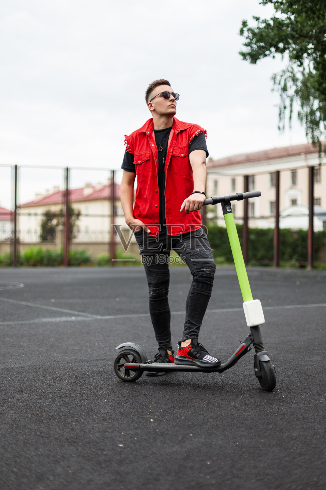 时尚的年轻男子穿着红色背心、黑色牛仔裤和运动鞋,戴着太阳镜,站在户外篮球场上,旁边是一辆现代电动滑板车。这个酷炫的嬉皮士正在骑完滑板车后放松。照片摄影图片