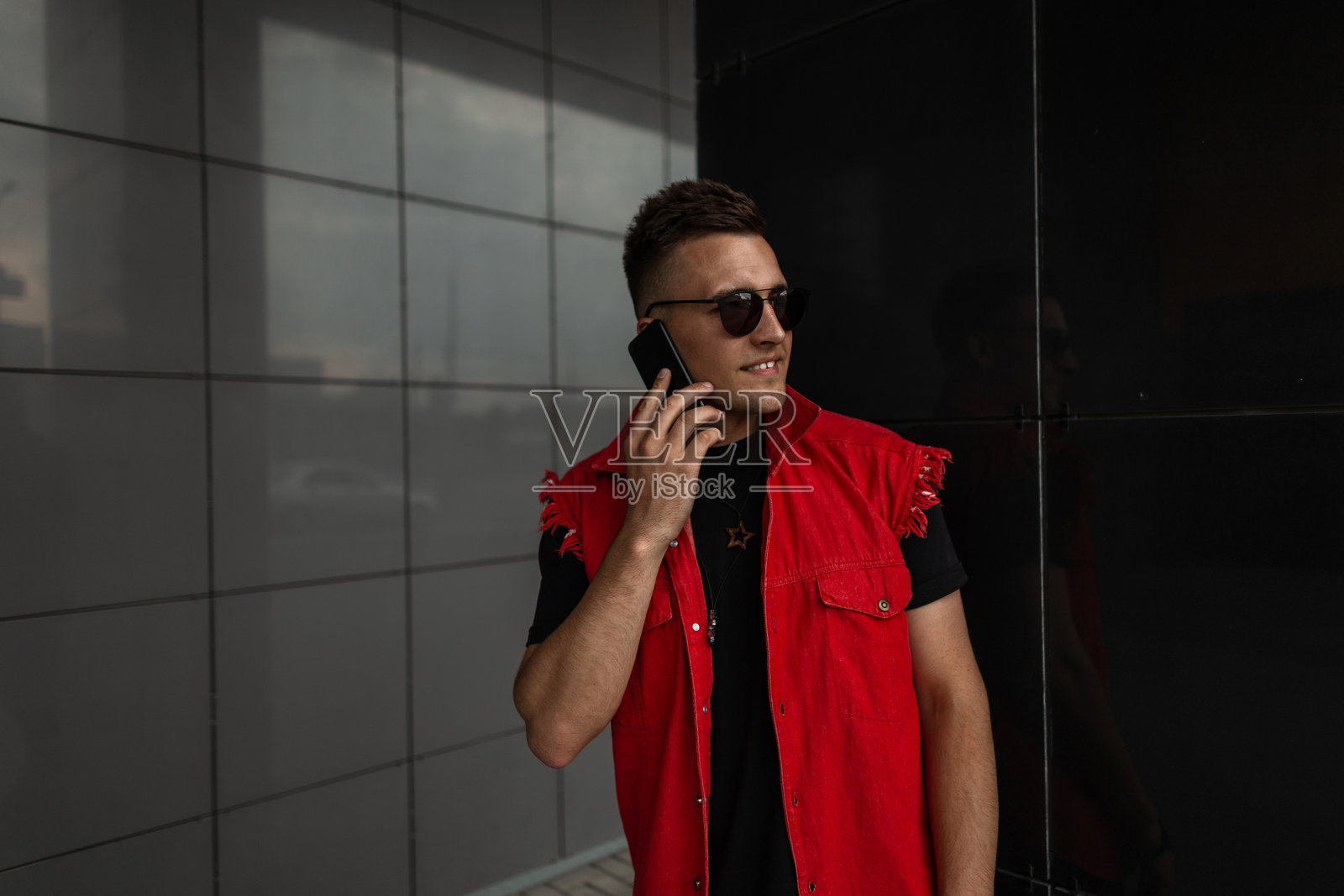 快乐帅气的年轻男子,带着美丽的微笑,佩戴着太阳镜,穿着时尚的红色牛仔服,站在黑色现代建筑旁边,正在打电话。可爱的快乐嬉皮士男孩在户外。照片摄影图片