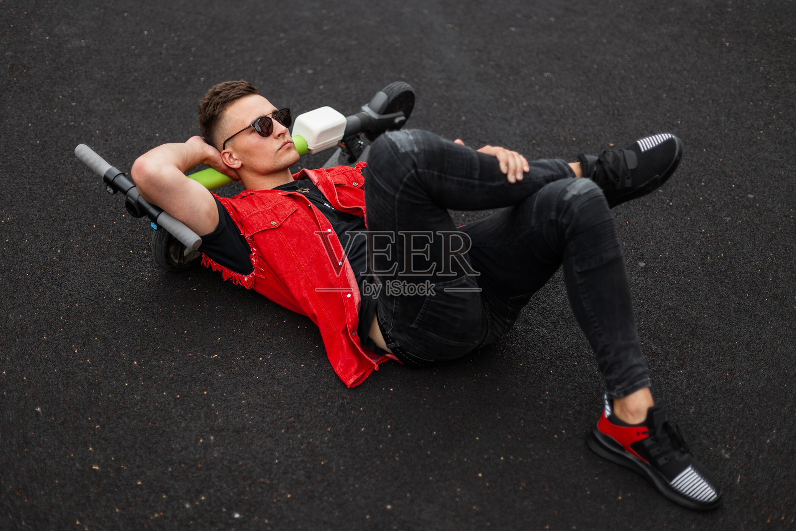 时尚的年轻男士,戴着太阳镜,穿着时尚的牛仔背心和黑色牛仔裤,脚踩运动鞋,躺在篮球场上的电动滑板车上休息。这个迷人的男孩是一名时尚模特。照片摄影图片