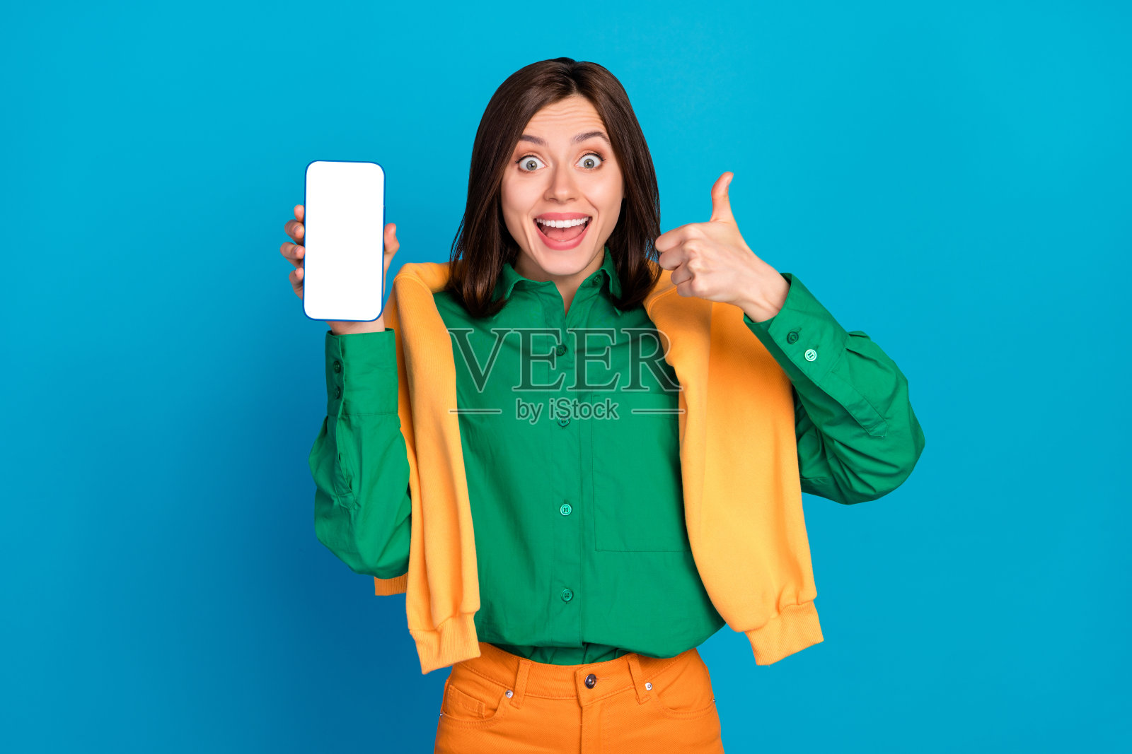 兴奋搞笑的女士手持空白电话显示屏,竖起大拇指表示反馈,背景为蓝色。照片摄影图片