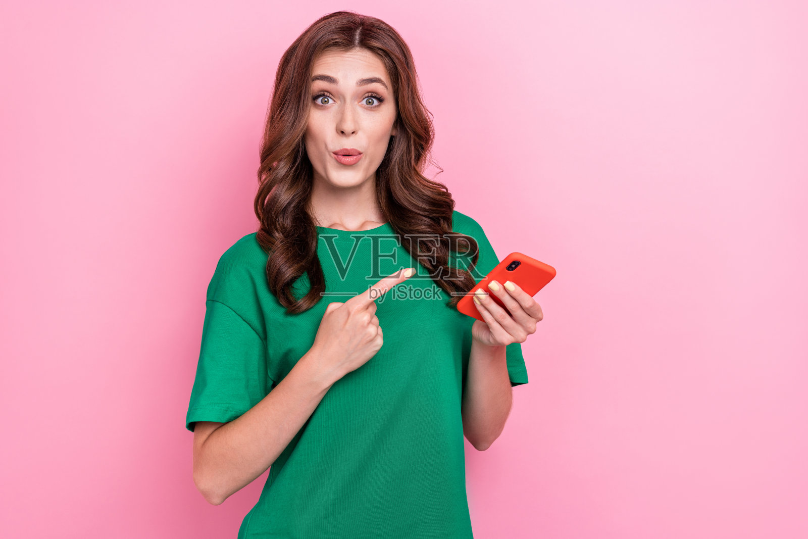 一位可爱迷人的女性,波浪卷发,穿着绿色T恤,面向智能手机,背景为粉色。照片摄影图片