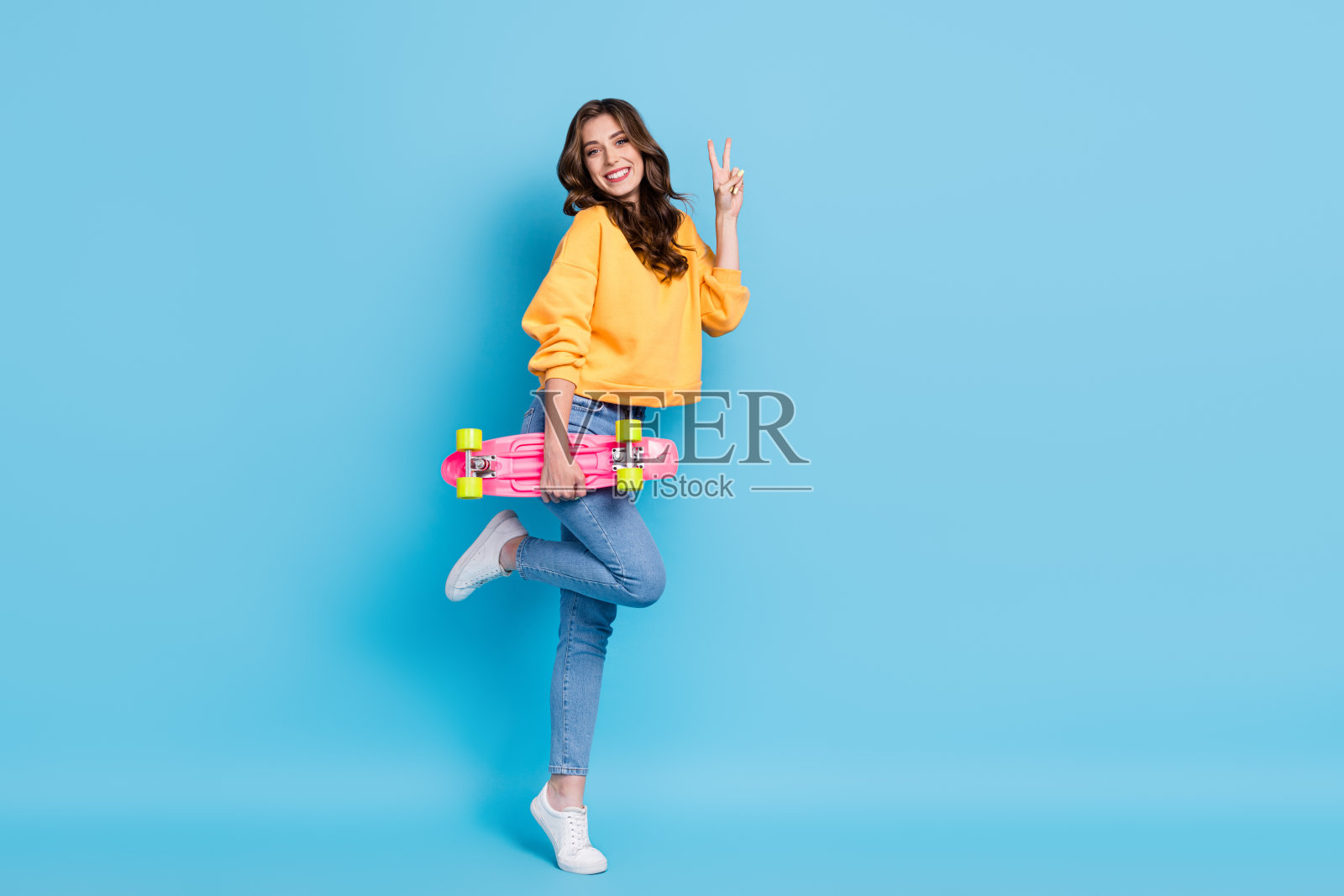 穿着黄色毛衣的酷炫漂亮女士,做出V字手势,手持长板,背景为空白的蓝色。照片摄影图片