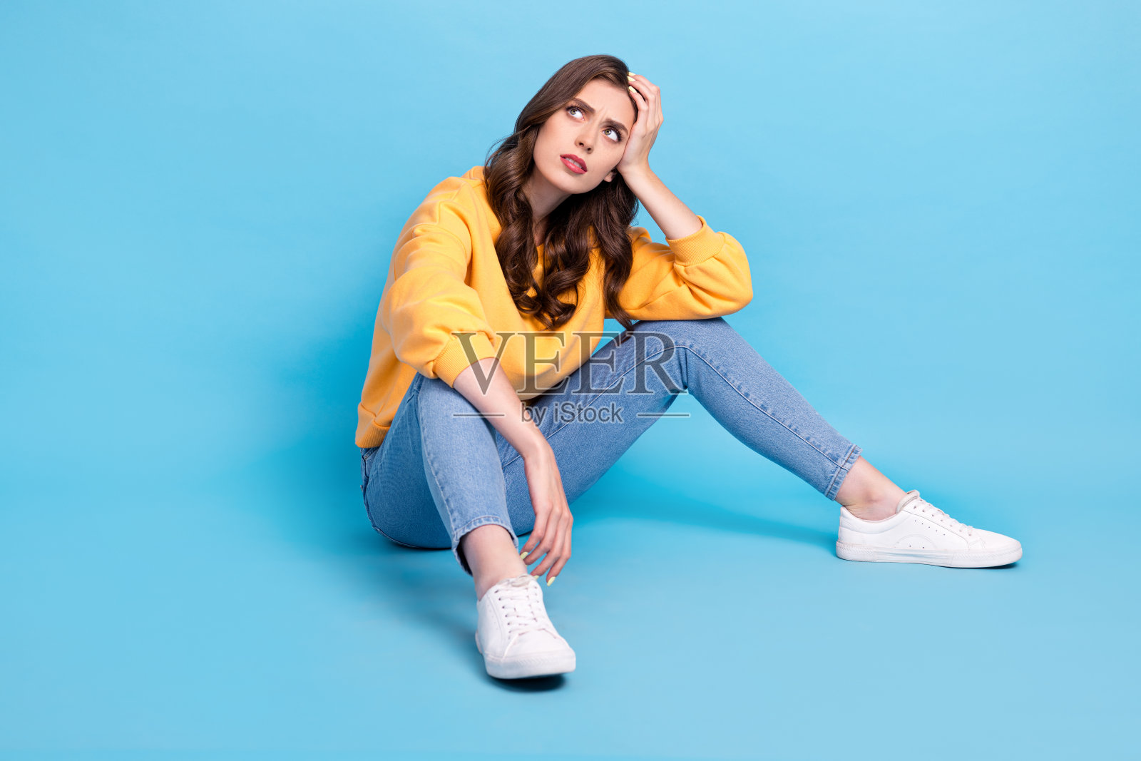穿着黄色毛衣的疑惑女士坐在地板上,目光空洞,背景为蓝色。照片摄影图片