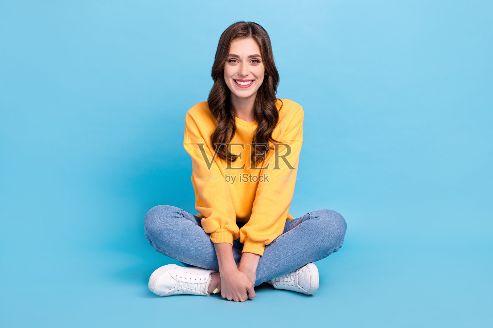 全身照,迷人的年轻女士坐在地板上,双腿交叉,放松休息,穿着时尚的黄色服装,背景为蓝色。照片摄影图片