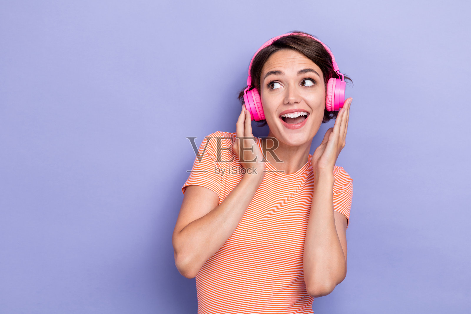穿着条纹时尚服装的兴奋快乐女士,面带微笑,注视空旷的空间,聆听音乐,背景为紫色。照片摄影图片